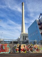 Person steht vor einer Wand mit bunter Graffiti vor einem hohen Industrie-Schornstein und einem blauen Gebäude