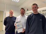 Drei Männer in Arbeitskleidung stehen in einem Raum, der mittlere hält ein Smart Meter