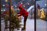 Frau in rotem Pullover und Mütze steht auf einem Hocker und schmückt einen Weihnachtsbaum mit Lichterketten im verschneiten Garten